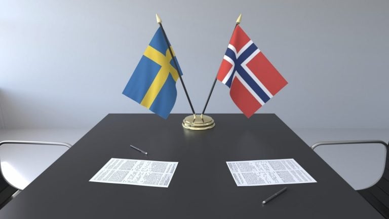 A rectangular table with two chairs is shown. On the table, there are two documents and two pens. Two small flags, one Swedish and one Norwegian, stand upright in a holder at the center of this distinctly Scandinavian setting.